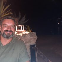 Smiling man with short dark hair and beard wearing a green polo, outdoors at night with plants and rocks behind him.